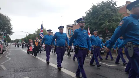 A band marches through Belfast, they are wearing dark blue trousers, lighter blue shirts and dark blue peaked caps. A man at the back of the procession wearing a black suit and orange sash is carrying a Union flag.