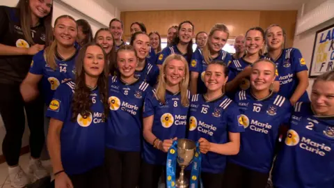 A school sports team of girls. All the girls are wearing blue jerseys. All the girls are smiling. Two girls at the front are holding a trophy.
