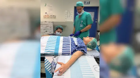 Emily Damari Family Emily Damari lays on an operating table wrapped in hospital blankets, with two health workers dressed in green behind her.