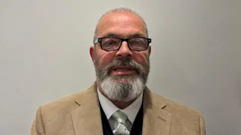 The image shows a man standing against a plain, light-coloured wall. He is facing towards the camera and is wearing black rectangular glasses and has short hair, a full greying beard and moustache. His clothing consists of a tan blazer worn over a dark sweater, with a white shirt underneath and a light-coloured tie.