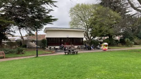 Daniel Mumby A cafe building with picnic tables outside set in a park with grass and trees surrounding it