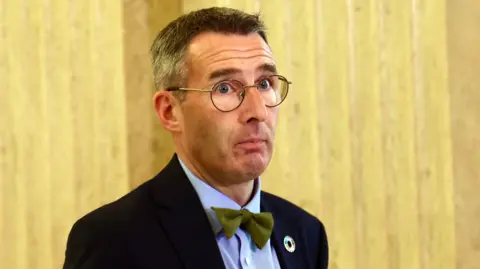 Pacemaker A man with grey and brown hair wearing circular glasses as well as a navy blazer, a green bow tie and a blue shirt stands in Stormont's Great Hall