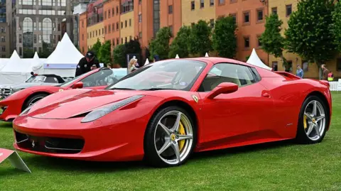 Getty Images A red ferrari sports car parks on a lawn with more another Ferrari and a Mercedes in the background as well as brick buildings in London