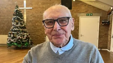 A close up of a man with black-rimmed prescription glasses and white hair, smiling straight to the camera. He's wearing a light grey v neck sweater and a pale blue polo shirt underneath. He's sitting in a community centre hall with wooden floors and brick walls, and behind him, to the left of the photo, there's a big green Christmas tree decorated with silver tinsel and white baubles. It has a silver star on the top of the tree.