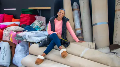 KDM PHOTOGRAPHY ST ALBANS A woman, Pia Honey, is sat on a pile of rolled up carpets in front of a pile of other carpets and cushions and she is smiling at the camera. Pia is wearing a pink top, black gilet, jeans and brown boots.