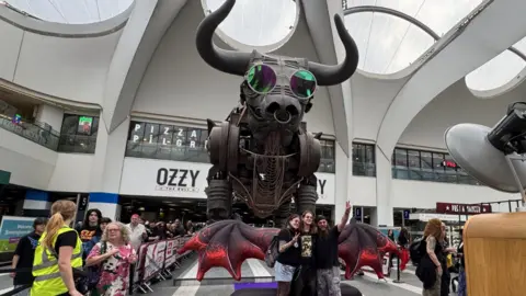 The large mechanical Ozzy the Bull statue is pictured in Birmingham New Street, wearing round pink glasses. An inflatable pair of red wings are positioned in front of him, and a group of p