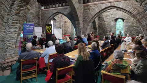 MANX LITFEST People at a writers workshop in a church building, Three authors are sitting at the front of the room.