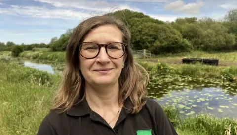 Claire Newill is standing in a field in front of a river. She is wearing a black shirt and glasses and has medium length brown hair.