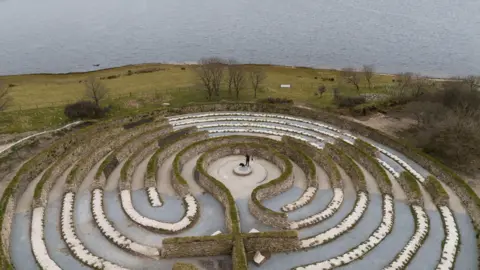 The picture shows an aerial drone shot of a circular labyrinth near a body of water. There are lots of paths separated by low hedges and walls. At the centre there is a small circular area with a man and a dog stood. 