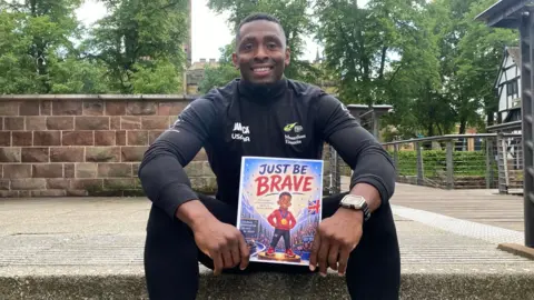 A black man in black running gear holding a children's book with the title Just Be Brave