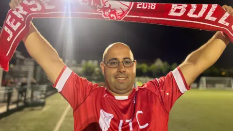 Joao holds a Jersey Bulls scarf over his head as he wears his red Jersey Bulls kit and is on the 3G pitch. He smiles at the camera and is wearing glasses.