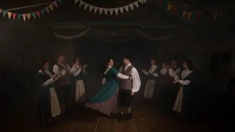Joann Randles A dark photo. There is a couple in the middle in traditional Welsh attire dancing, the woman holds on to the mans waist. She has a tartan underskirt, with a blue dress on the top, and you can see her lower arms covered in a white sleeved undershirt. The man has a white shirt on and a tartan vest. He has black trousers and khaki socks rolled up with black shoes. There are three people to the left of them clapping, who have the same attire on - one man and two women. On the right there are two women and a man clapping. There are triangular banners strung above the ceiling at the top of the image.