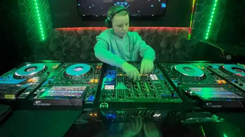 Kerry O'Kane Fionn is seen in a grey jumper with a pair of headphones on.
He is at a DJ booth with a number of green lights behind him.
He is mixin and staring at the decks.