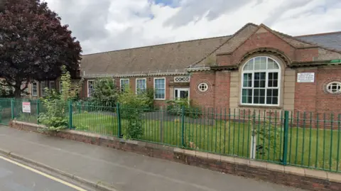 An old-fashioned single storey red brick building under a steep tile roof. There is  green metal fencing and grass in front of it
