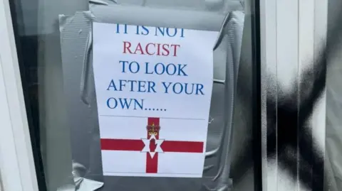 Sign taped to a model   speechmaking  'It is not racist to look   aft  your ain  .....' with Northern Ireland emblem  beneath   