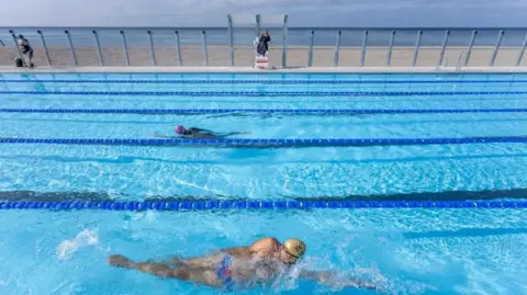 People swimming in a swimming pool with a lifeguard on duty. In the background is Brighton beach.