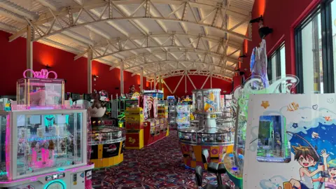 Andrew Turner/BBC The inside of a small building painted red which has an arched white roof. Multiple bright arcade games are dotted around the room.