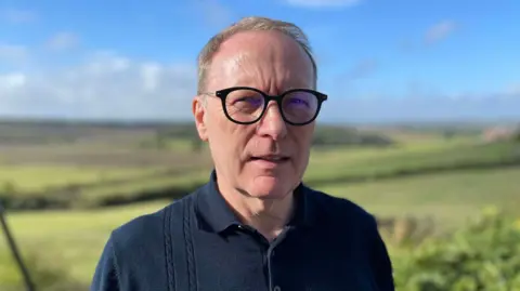 A man in his early 60s wearing glasses and a navy top standing in front of a valley with blue skies and rolling green fields