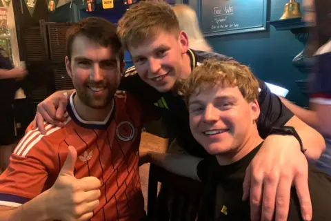 Scotland fans (from left) Ryan Kerr and Matthew and David Smith. They are sitting in a pub. Ryan has his thumb raised and Matthew is standing with his arms round the other two.