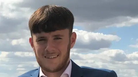 A closeup photo of a young man dressed in a navy blazer and white shirt. He has a long brown fringe and a short beard. 