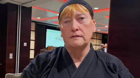 Roisin Pitman is wearing a black martial arts outfit. She has red hair.