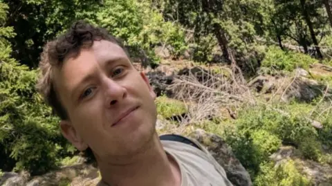 A man with short brown hair which is slightly curly on top taking a selfie surrounded by rocks, trees and general greenery. He is wearing a beige top and a black backpack.