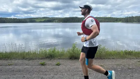 A man is running alongside a large open lake with green hills and pine forests on the horizon.