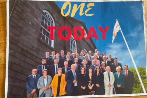 A card with the Guernsey States building a a group photo of the current deputies on it. It says 'ONE TODAY' at the top