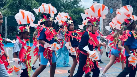 Children are wearing red costumes and dancing within a school's ground.