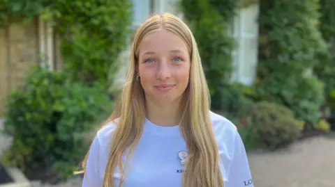 Person with long light-colored hair stands in front of a house entrance covered in greenery, wearing a white “King’s Baton Relay Glasgow 2026” shirt; the background shows windows, foliage, and a garden setting.