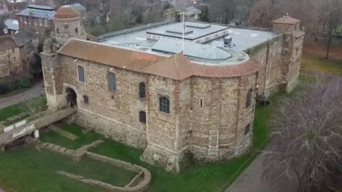 An aerial view of Colchester Castle, taken from a drone. The castle is made of yellow stone walls with a mostly flat roof. There is grass surrounding the castle with exposed foundations, and there is a drawbridge going into the main entrance of the castle.