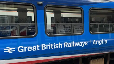 The side view of a railway carriage at Liverpool Street Station. The train is painted in bright blue with Great British Railways Anglia written in white letters under its windows. Beneath that on the edge of the body of the carriage is a line of red and white, decreasing to a point on the right.