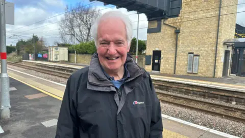 Jessica Bayley/BBC A man with white hair smiling wearing a waterproof coat, stood on a train platform with the train tracks behind him.