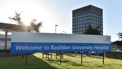 Exterior view of a hospital building. In the foreground is a sign which reads "Welcome to Basildon University Hospital". In the background is a tall grey coloured block with windows. 