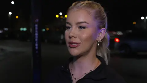 Katie Moys has blonde hair tied back in a pony tail. She is wearing a black top and has a silver necklace and earrings. She is standing on a street at night.