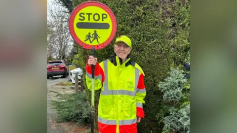 Reg Bown Reg dressed in a high-vis coat - he is holding a lollipop sign reading 'STOP' with the highway code symbol for children/ school below.