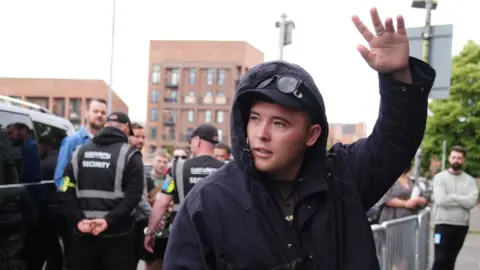 Liam Ó hAnnaidh, who is wearing a dark raincoat with the hood up over a black cap and sunglasses, waves to crowds outside Glasgow's O2 Academy. Behind him are his security team standing in front of a dark vehicle.