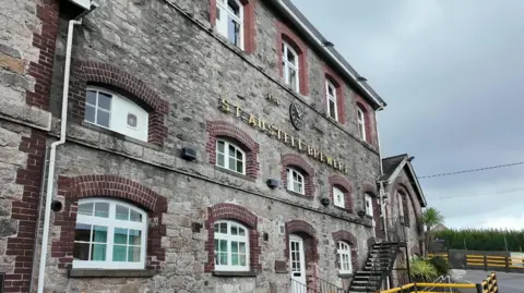 BBC The picture shows a stone building with multiple windows with a sign reading St Austell Brewery attached to the front wall. The building appears to have three floors, with the ground floor having white-framed windows and a door. There is a plaque on the wall near the door, and yellow and black railings line the pathway in front of the building. 