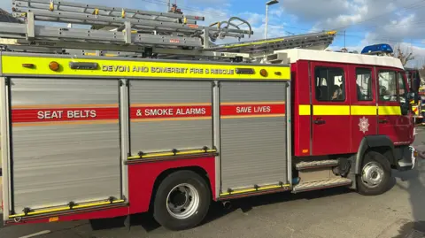 A Devon and Somerset Fire Service engine parked up on a road. It is red with metal shutters along the side. It has the fire service's badge on the side.