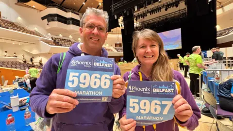 A man with grey hair and glasses, and a woman with long brown hair, both wearing purple hooded sweatshirts, stand indoors at a large event hall holding Belfast City Marathon 2026 race bibs. The bibs read “Phoenix Energy Belfast City Marathon 2026,” with numbers 5966 and 5967 labelled “MARATHON”; rows of seating, event staff in green shirts, and a stage with lighting are visible in the background.
