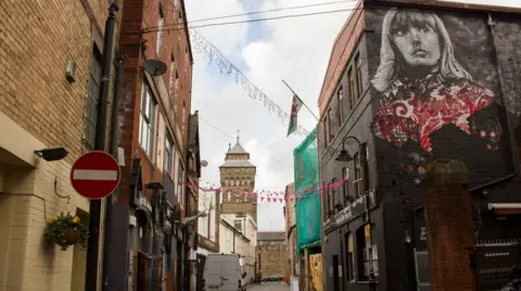 Jeremy Segrott/WikiCommons A narrow street with buildings on either side, bunting hanging above and Cardiff Castle in the distance.