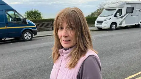 BBC/Seb Cheer A woman, Julie Anne, looks at the camera with a neutral expression on her face. Behind her, two campervans are parked at the side of a pavement.