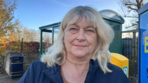 Jacqui Clark looks directly at the camera, she has blonde mid-length hair with a fringe and wears a navy blue waterproof jacket. She stands in front of a bus stop, which is blurred in the background.