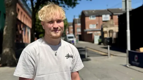 BBC / Hazel Shearing Sam stands in the sunshine at Nottingham. He wears a grey t-shirt and a silver necklace.