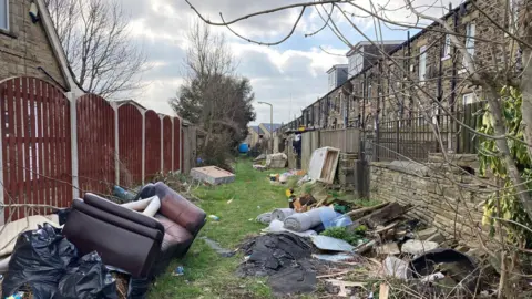 BBC A back alleyway lined with discarded rubbish including a brown leather settee and rolls of blue/grey carpet.  