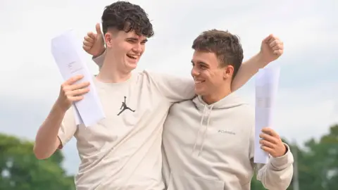 Two male students celebrate outdoors, with their arms around each other, each holding their results. The student on the right is giving a thumbs up. Both have short brown hair and are dressed casually in light-coloured clothing. 