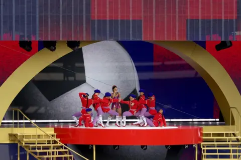 Getty Images Doja Cat performs during the halftime show at the FIFA Club World Cup 2025 Final match between Chelsea FC and Paris Saint-Germain at MetLife Stadium wearing a short purple and red dress. She is surrounded by dancers in red outfits and purple tights.