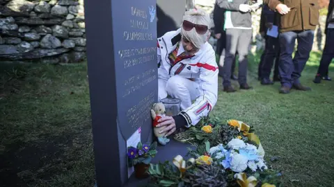 Getty Images Gina Campbell places Mr Whoppit - her father's teddy beat mascot - next to his headstone. Flowers are laid alongside. Gina has a sunglasses raised on to the top of her head and is wearing a white jacket and trousers. Onlookers can be seen in the background.