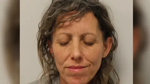 Police mug shot of Kara Alexander, a woman with blonde and grey curly hair, with her eyes closed 