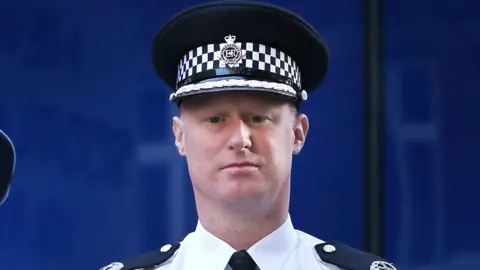 Steve Rodhouse, a clean-shaven Caucasian male, wearing a white police shirt with police issue epaulettes, a black tie and police hat.